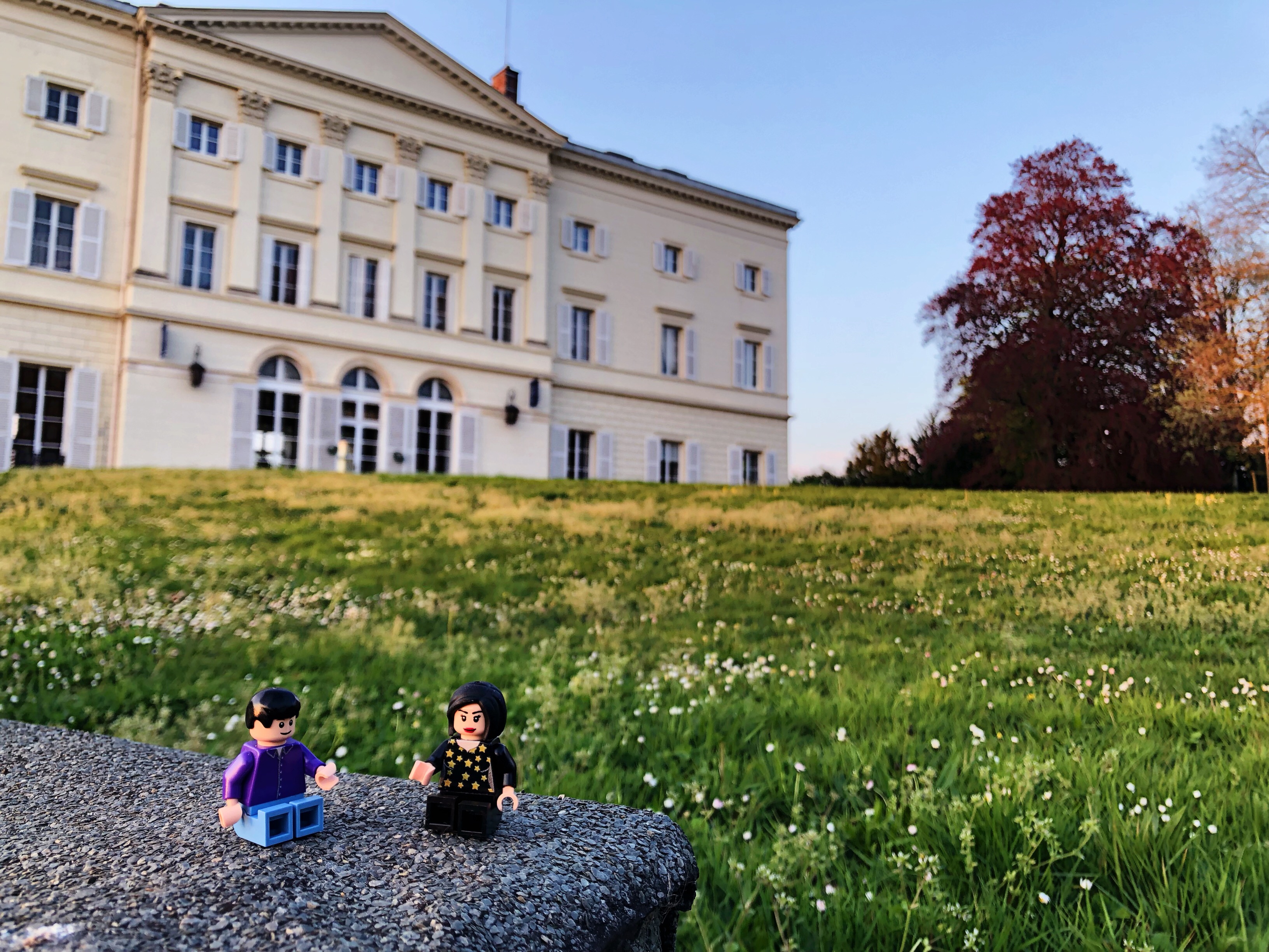 A lego man and woman sat together on stone in the grass in front of the HEC Chateau, which features in the background along trees of varying colours