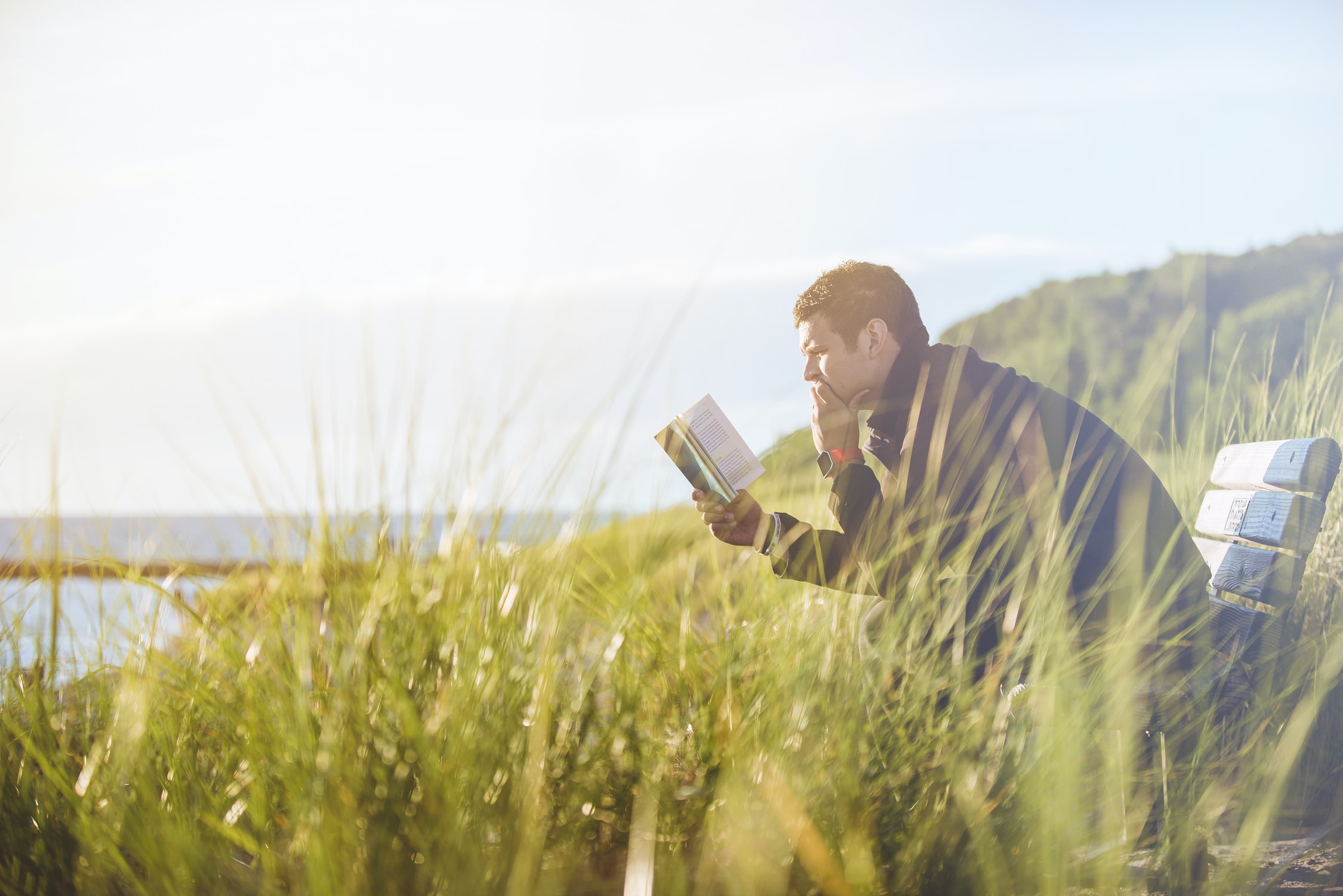 Man reading a book by the seaside, with alone with lots of green grass and mountains in the background